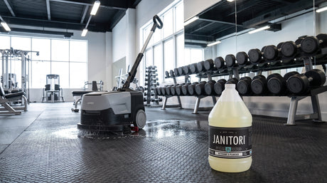 Compact commercial auto scrubber cleaning a black rubber gym floor beside weight racks with a JANITORI Floor Cleaner No.61 jug